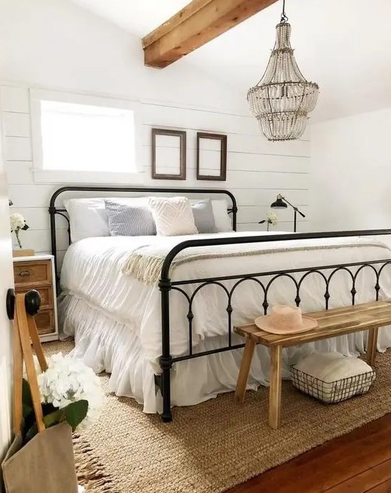 a neutral farmhouse bedroom with a metal bed, wooden nightstands and a bench, a crystal chandelier and neutral bedding