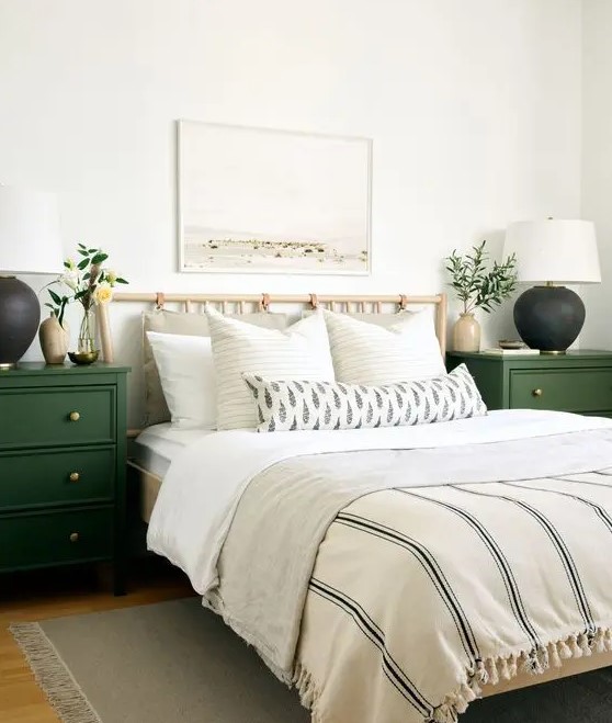 a neutral farmhouse bedroom with a wooden bed with neutral bedding, green nightstands, two-tone lamps and greenery