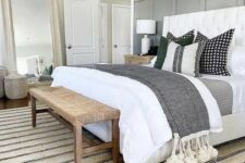 a neutral modern farmhouse bedroom with a grey paneled wall, a white bed with neutral bedding, a woven bench, a mirror and a rug
