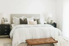 a neutral modern farmhouse bedroom with a grey upholstered bed with neutral bedding, a leather bench, black nightstands that are dressers
