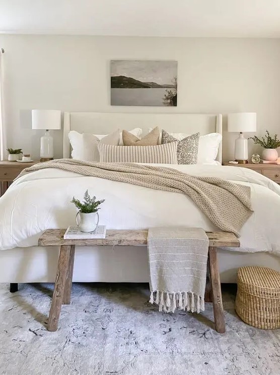 a neutral modern farmhouse bedroom with an upholstered bed with neutral bedding, a stained bench, stained nightstands, a basket and some decor