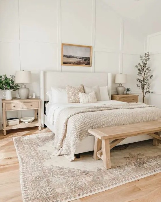 a neutral modern farmhouse bedroom with paneled walls, a neutral upholstered bed with neutral bedding, a wooden bench, wooden nightstands and greenery