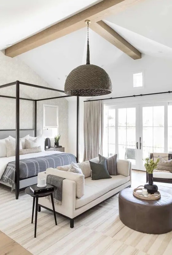 a neutral modern farmhouse bedroom with wooden beams, a blakc frame bed with neutral bedding, creamy seating furniture and a woven pendant lamp