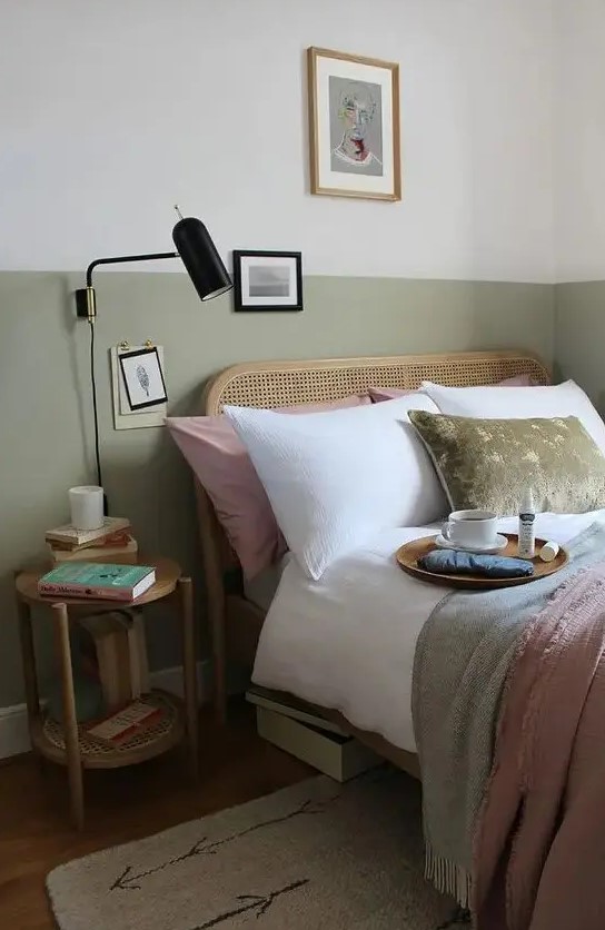 a pretty and welcoming bedroom with white and olive green walls, a rattan bed and neutral and green bedding, a wooden nightstand and a black sconce