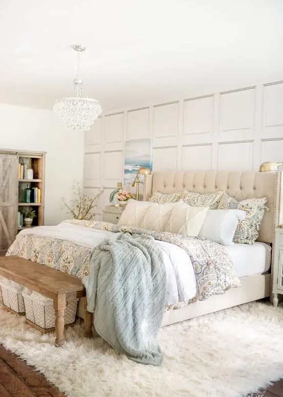 a pretty coastal farmhouse bedroom with a paneled wall, an upholstered bed, a wooden bench, a chic chandelier and a a fux rug