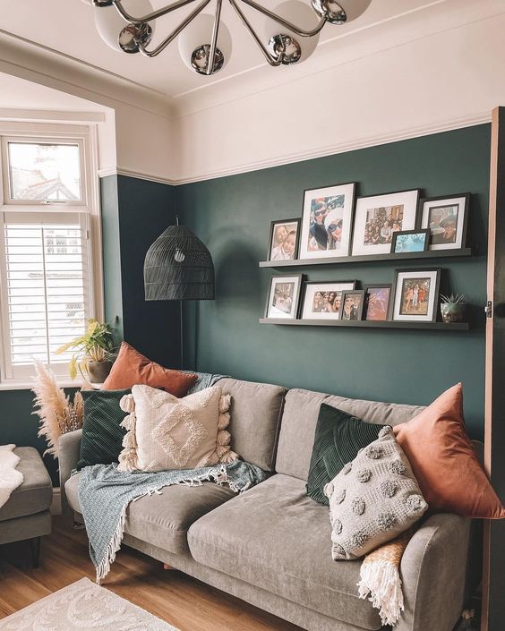 a pretty living room with dark green walls, a grey sofa with pillows, a ledge gallery wall and a black pendant lamp