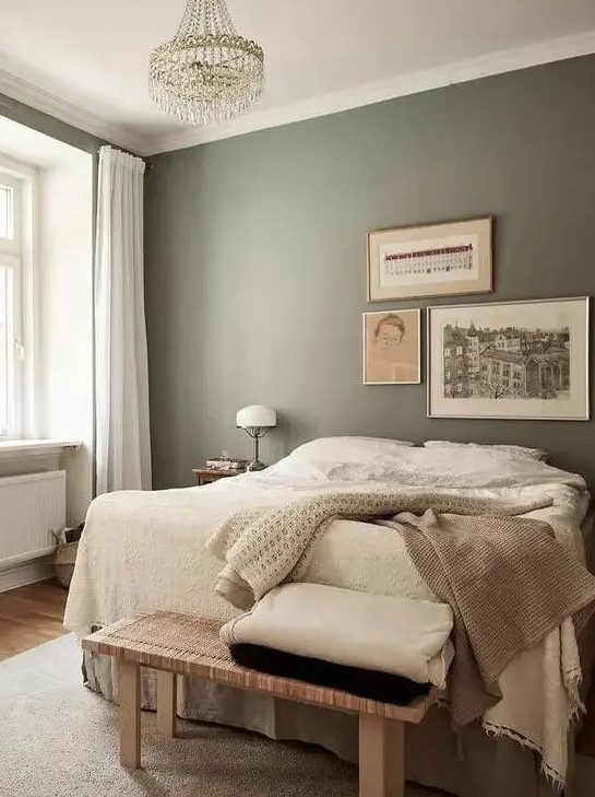 a relaxing bedroom with olive green walls, a bed with neutral layered bedding, a woven bench, a crystal chandelier and some art