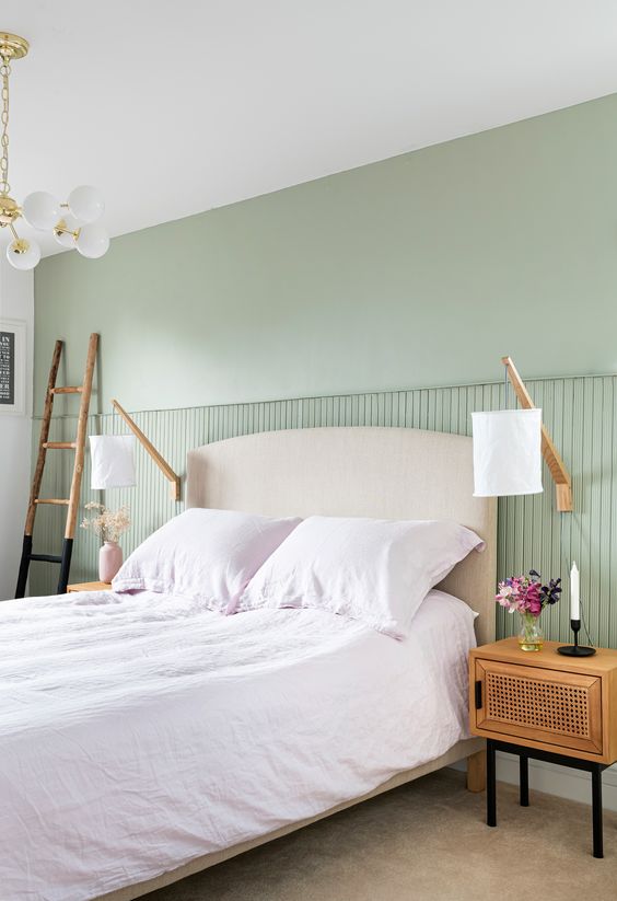 a serene bedroom with a light green accent wall, a neutral upholstered bed, cane nightstands, sconces and a ladder