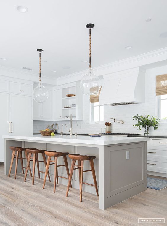 a serene modern farmhouse kitchen with white cabinets, a dove grey kitchen island, pendant lamps, wooden stools and greenery