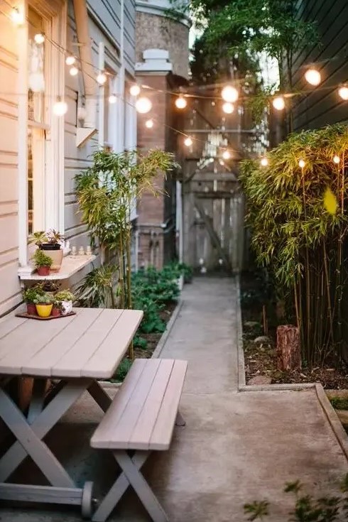 a side yard with a concrete path, a wooden dining set, bamboo and greenery and string lights