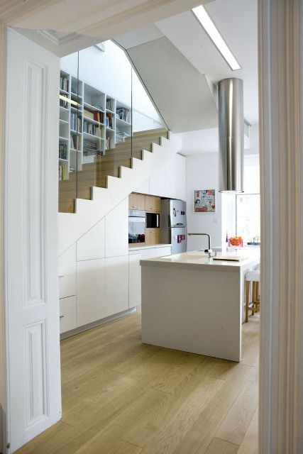 a sleek white kitchen built under the stairs, with appliance,s a kitchen island, a hood over it and some tall stools