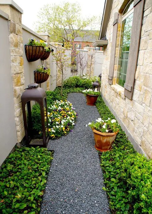 a stylish and manicured side yard with a gravel path, greenery, blooms and pots with blooms and an oversized lantern