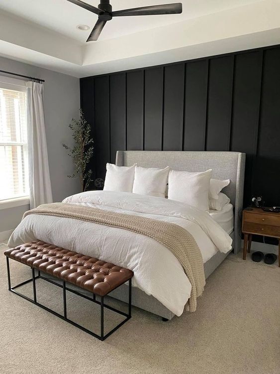 a stylish modern farmhouse bedroom with a black paneled wall, a neutral bed with neutral bedding, a brown leather bench and a stained nightstand