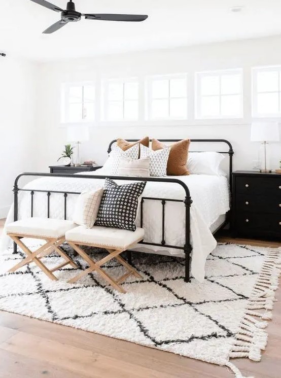 a stylish modern farmhouse bedroom with a row of windows, a wrought bed with neutral bedding, stools and black nightstands