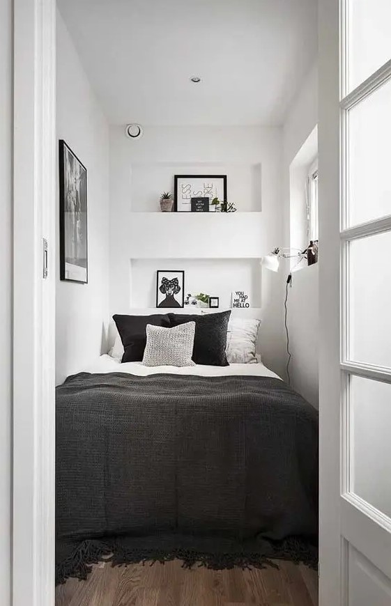 a tiny bedroom in white, with only a bed squeezed in, black and white bedding, niche shelves with decor, an artwork and a sconce