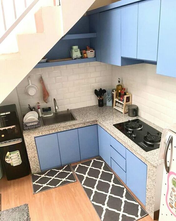 a tiny blue kitchen built under the staircase, with a subway tile backsplash and terrazzo countertops and black rugs