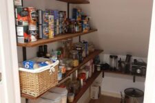 a tiny under stairs pantry with stained open shelves, a basket, plastic containers and a lot of food and stuff stored