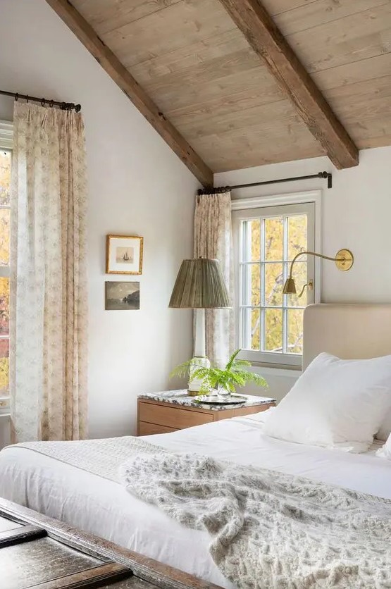a welcoming modern farmhouse bedroom with a wooden ceiling, a neutral upholstered bed with neutral bedding, stained nightstands, printed textiles