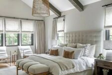 a welcoming modern farmhouse bedroom with wooden beams, a neutral upholstered bed with neutral bedding, a bench and some chairs, stained nightstands