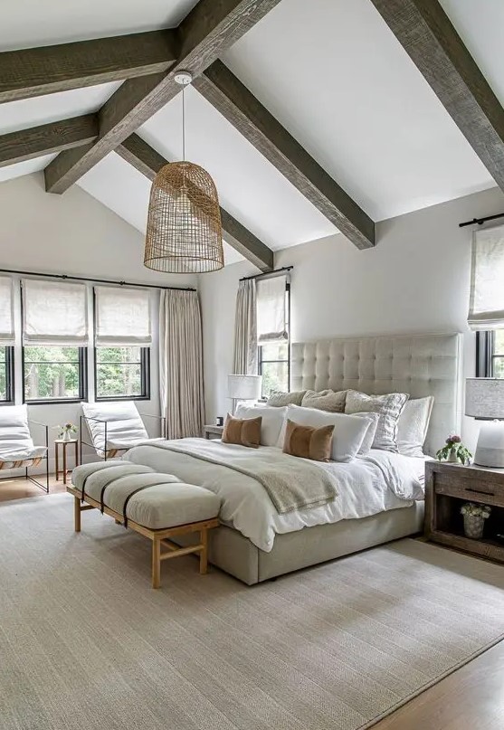 a welcoming modern farmhouse bedroom with wooden beams, a neutral upholstered bed with neutral bedding, a bench and some chairs, stained nightstands