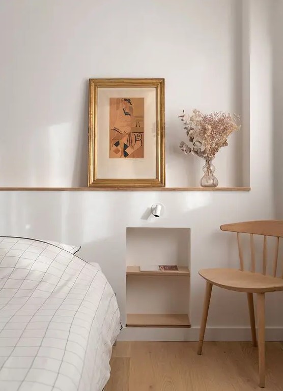 a welcoming neutral bedroom with a bed and neutral bedding, a niche with shelves, some art and a stained chair