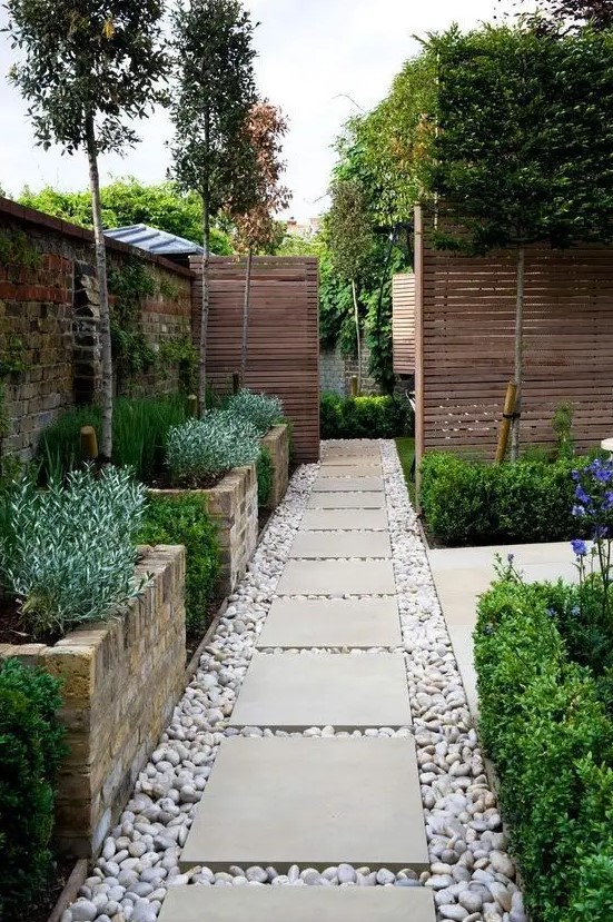 a welcoming pathway with large brick flower beds with greenery and pebbles on the ground with large tiles