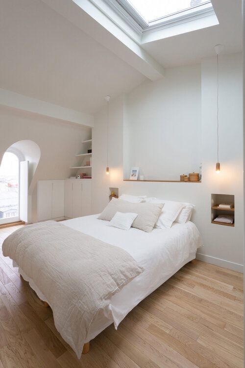 a white attic bedroom with a skylight, bed with neutral bedding, a niche as a display shelf and some niches as nightstands, some attic shelves