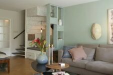 an airy living room with light green walls, a grey sectional, some coffee tables, a grey rug and cool decor