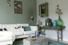 an eclectic living room with a grey and green wall, a white sectional with pillows, a green shabby chic console table and a suitcase as a table