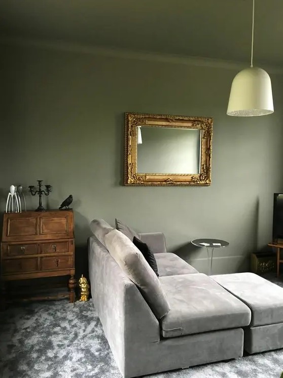 an elegant living room with olive green walls and a ceiling, a grey sectional, a stained storage unit, a mirror in a frame and a pendant lamp