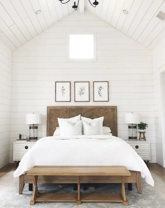 an elegant modern farmhouse bedroom with a bed with a wooden headboard, a stained bench and white nightstands, a gallery wall