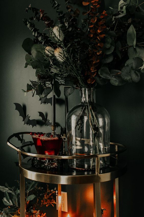 a gorgeous spooky bar cart with dark bloody drinks, a bottle with moody and dark blooms and leaves and bats on the wall
