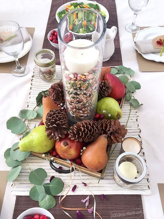 a fall centerpiece with pears, pinecones, a glass with corn and a candle plus greenery around can be easily made
