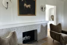 16 a refined and chic living room with a fluted fireplace, taupe chairs, an artwork and sconces, a rug