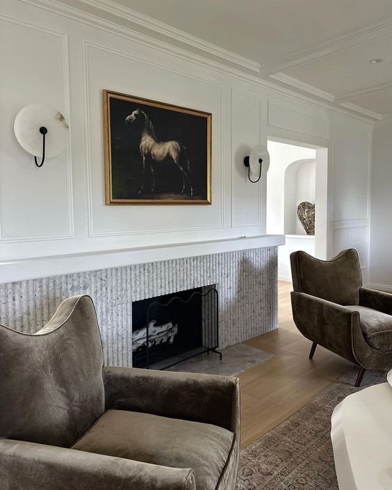 a refined and chic living room with a fluted fireplace, taupe chairs, an artwork and sconces, a rug