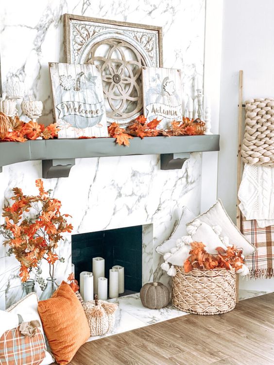 a fall mantel with candles, bright faxu leaves, pumpkins, a basket with pillows and branches and branches in a bottle
