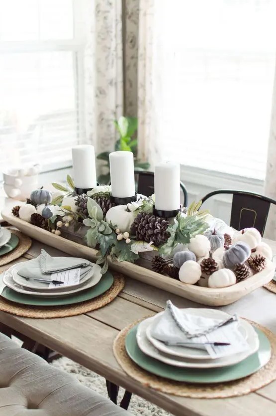 a stylish fall centerpiece of a white bowl with pinecones and mini pumpkins and pillar candles in the center