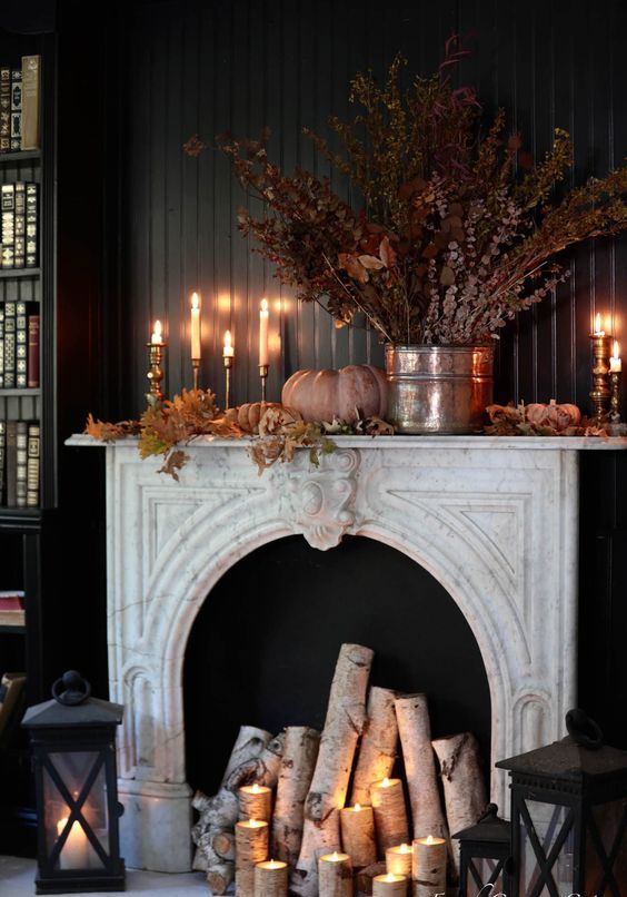 an elegant and refined fall mantel with firewood, pillar candles, faux leaves, a pumpkin and a dried leaf arrangement