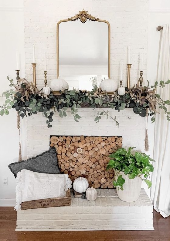 an elegant rustic fall mantel with greenery, white pumpkins, brass candlesticks and a mirror, a wood slice screen in the fireplace is chic
