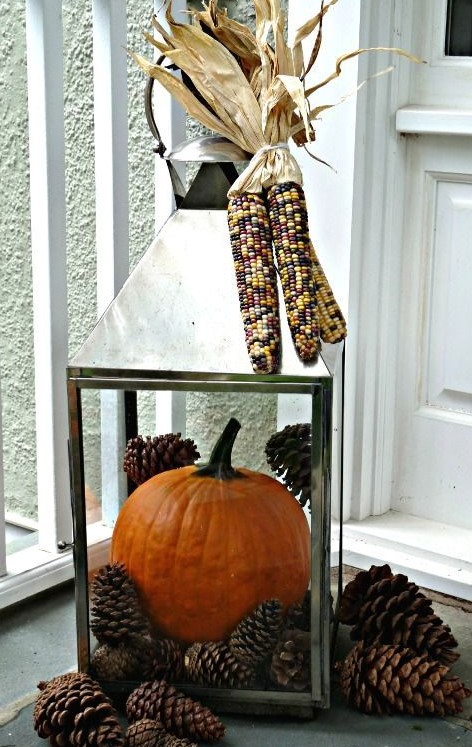 a large lantern filled with pinecones and a large orange pumpkin plus corn on top for a rustic feel