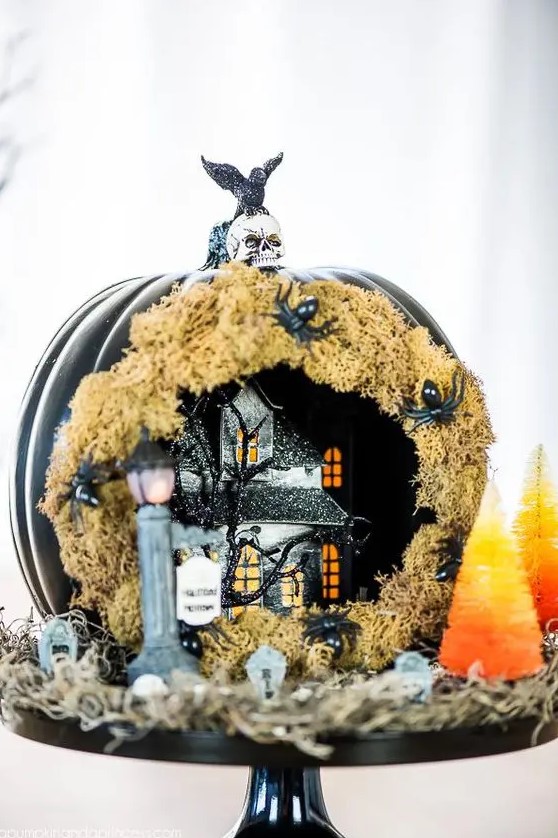 a Halloween diorama with a black pumpkin covered with moss and spiders, a black house with lights and a tree and some tombstones