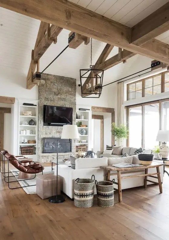 a barn living room with wooden beams, white planked walls, a fireplace clad with stone, neutral seating furniture, leather chairs and baskets