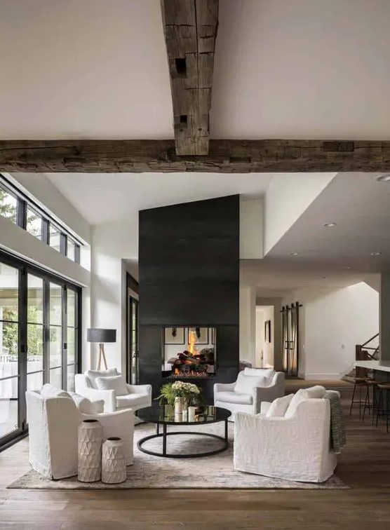 a beautiful modern farmhouse living room with reclaimed wooden beams, a large double-sided fireplace, white seating furniture and a coffee table