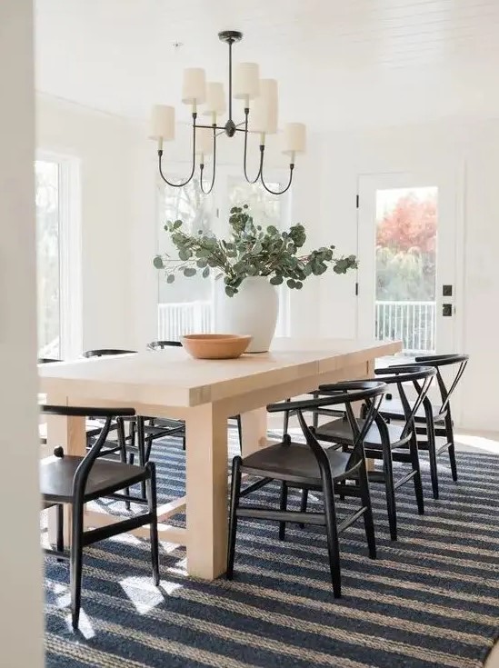 a cool modern farmhouse dining room with a light stained table, black wishbone chairs, a chandelier and a striped rug