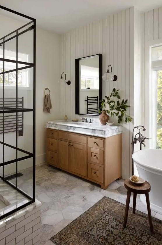 a cozy modern farmhouse bathroom with shiplap walls, a shower space, a vanity and a mirror, a tub, a wooden stool and a boho rug