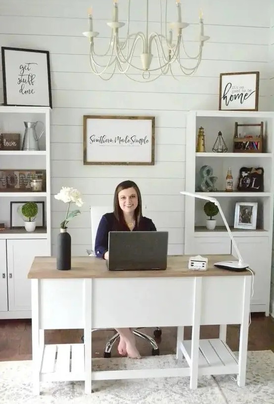 a cute white modern country home office with a planked wall, built in storage units, a large desk and a neutral chandelier