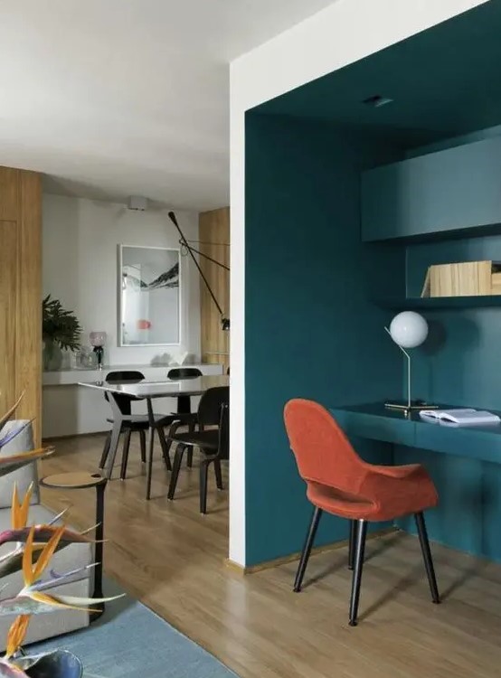 a deep teal niche with built-in shelves and a desk, an orange chair, a table lamp and built-in lights is great for working
