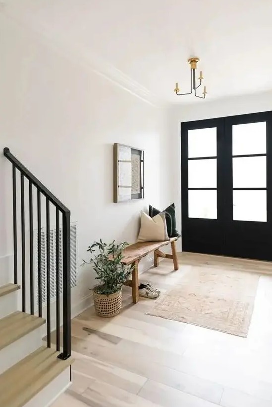 a farmhouse entryway with black doors, a stained bench and black and white pillows, a printed rug and a potted plant