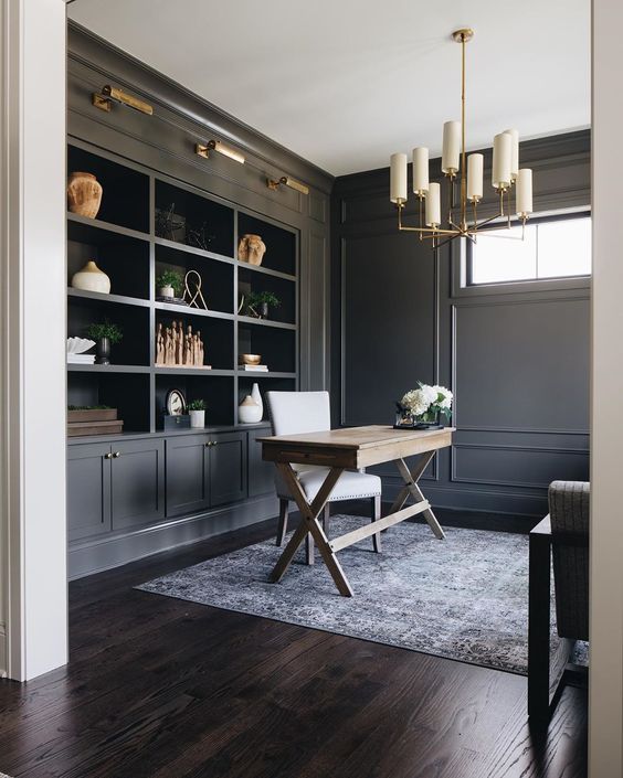 a graphite grey modern farmhouse home office with built-in storage units, a trestle desk, a white chair and a chic chandelier