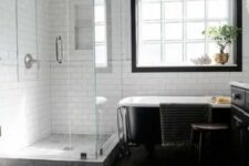 a laconic monochromatic farmhouse bathroom with a dark wooden floor, white subway tiles, a black clawfoot bathtub and a window